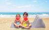 Dwoje dzieci siedzi na macie plażowej, każde trzyma kawałek arbuza, a w tle widać ocean i czyste niebo.